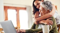 Young woman hugging her mother who is typing on a laptop