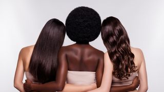 photo shows three woman from the back as they stand in a line with their arms around one another. the woman on the left has tan skin and long, dark brown hair. The center woman has black skin and an afro. The woman on the right has pale skin and long, wavy brown hair
