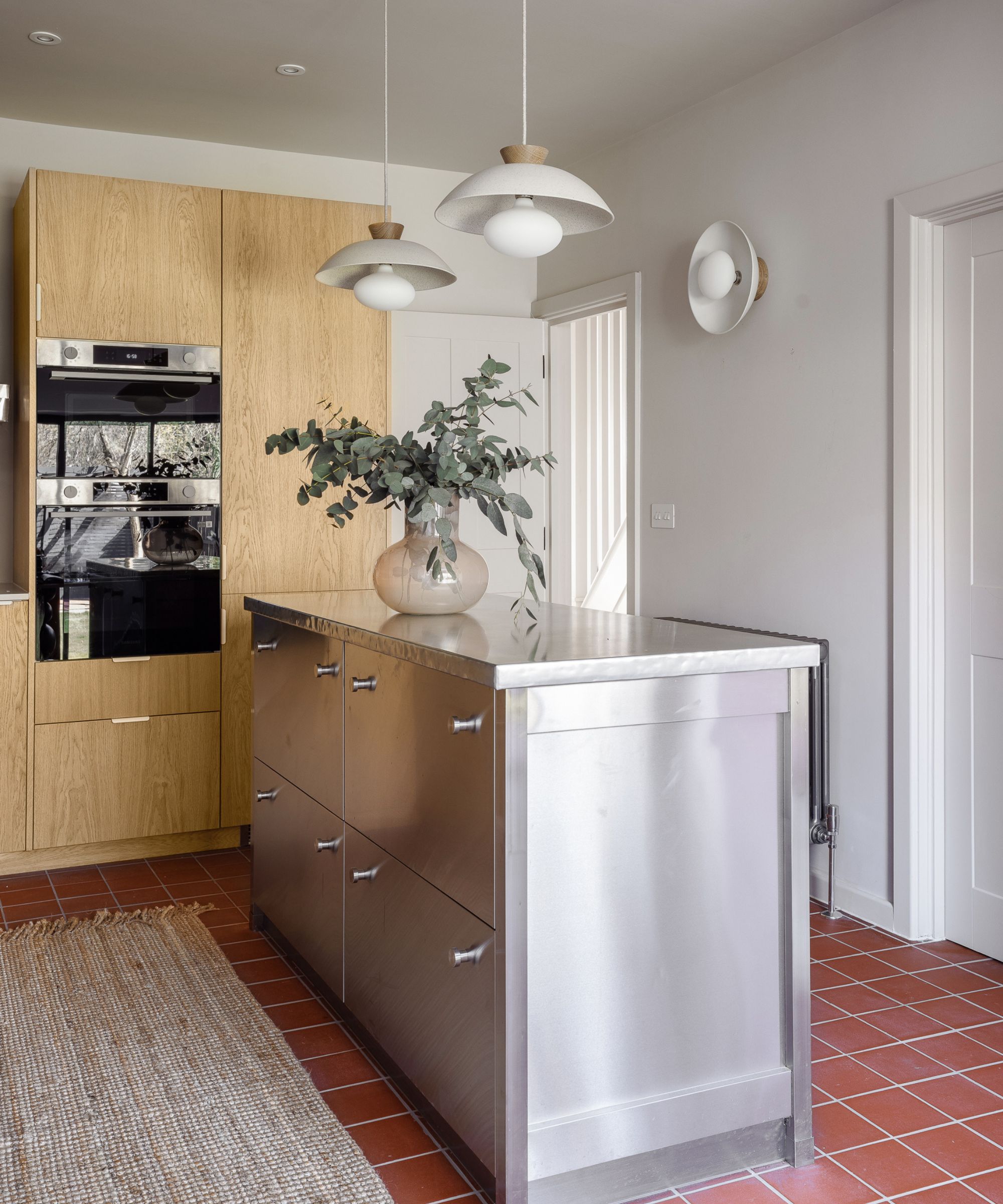 A bright kitchen featuring a stainless steel island with minimalist pulls on a terracotta tile floor. Two white mid-century modern pendant lights hang above, with oak wood cabinets and a built-in oven in the background.