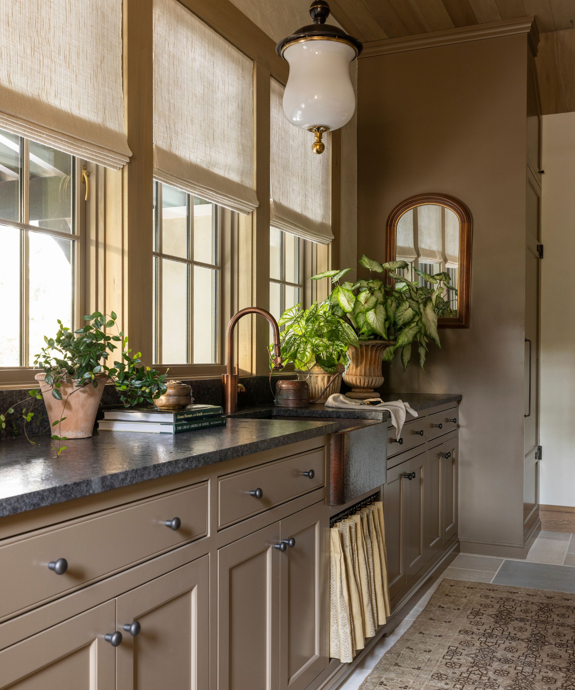 Mudroom with cabinetry and walls painted in Farrow &amp;amp; Ball's Broccoli brown, three large windows, a dark marble countertop, a sink with a skirted cupboard underneath, and plants in rustic pots.