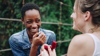 A woman holds up a red engagement ring box in front of another woman