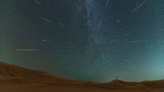 A long exposure perseid meteor shower image of the night sky showing tens of perseid meteors streaking through the sky as long white lines against a backdrop of stars. In the distance a person stands with arms outstretched to the sky.