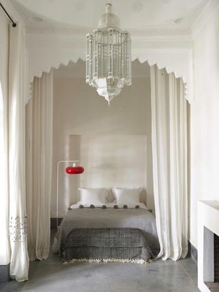 A hotel bedroom with a bed, a grey ottoman, a floor lamp, a fireplace, white curtains and a white chandelier.