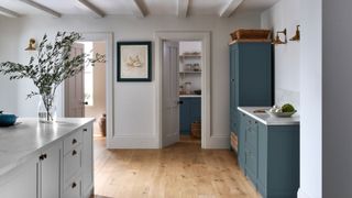 A country kitchen with sage green and teal blue shaker cabinets, brass knob handles, white marble worktops, oak wood floors, painted beams and an open pantry doorway.