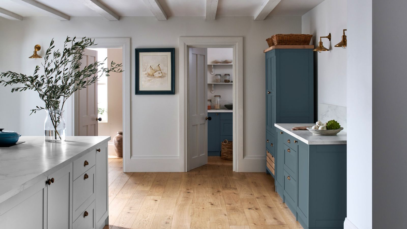 A  country kitchen with sage green and teal blue shaker cabinets, brass knob handles, white marble worktops, oak wood floors, painted beams and an open pantry doorway.