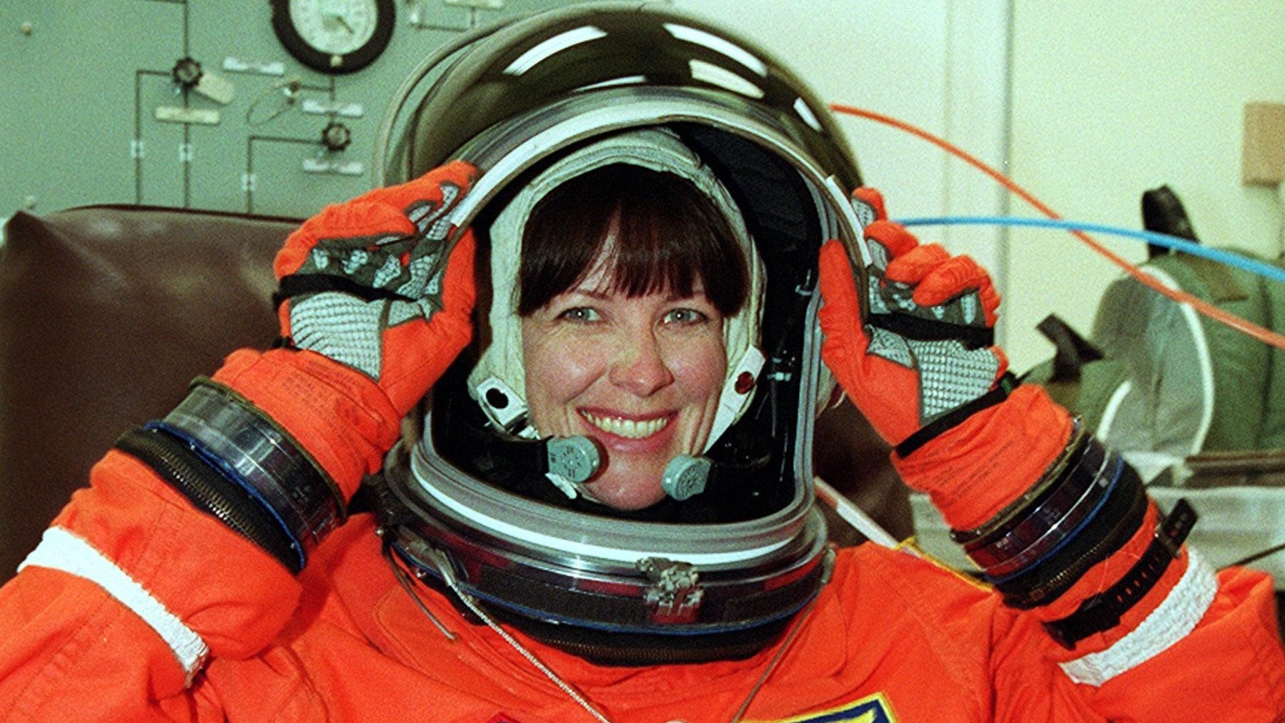 nasa astronaut janet kavandi wearing a helmet with open visor and an orange launching spacesuit. she smiles and grasps the visor of the helmet with both hands. behind her is medical equipment