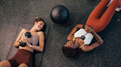 Two woman lay opposite each other on a gym mat with a weight in hand after doing the best gym workouts 