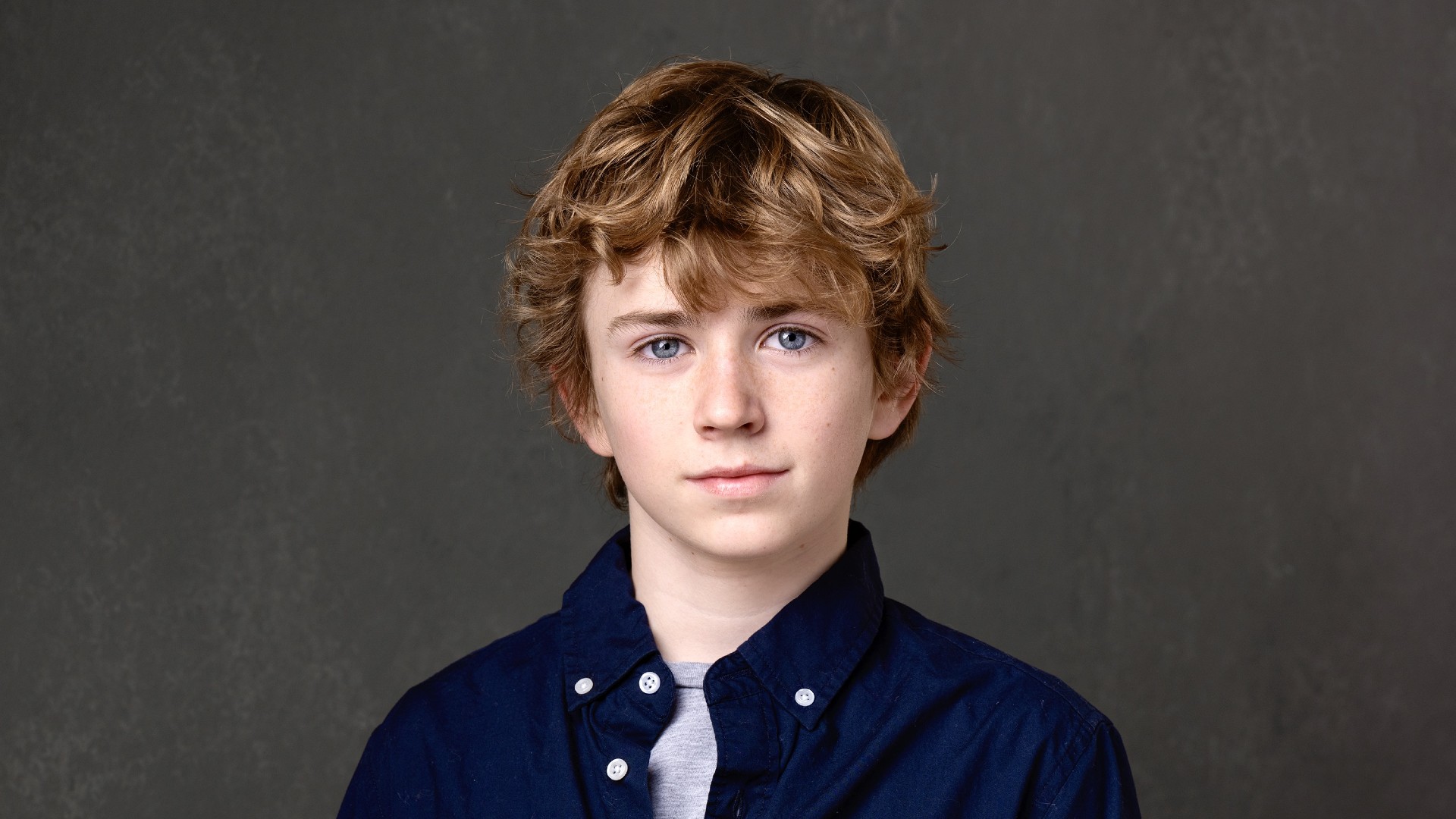 A professional headshot of young actor Walker Scobell. He has short, curly blond hair, slightly pale skin, and blue eyes. He is wearing a dark blue shirt with a light gray t-shirt undearneath.