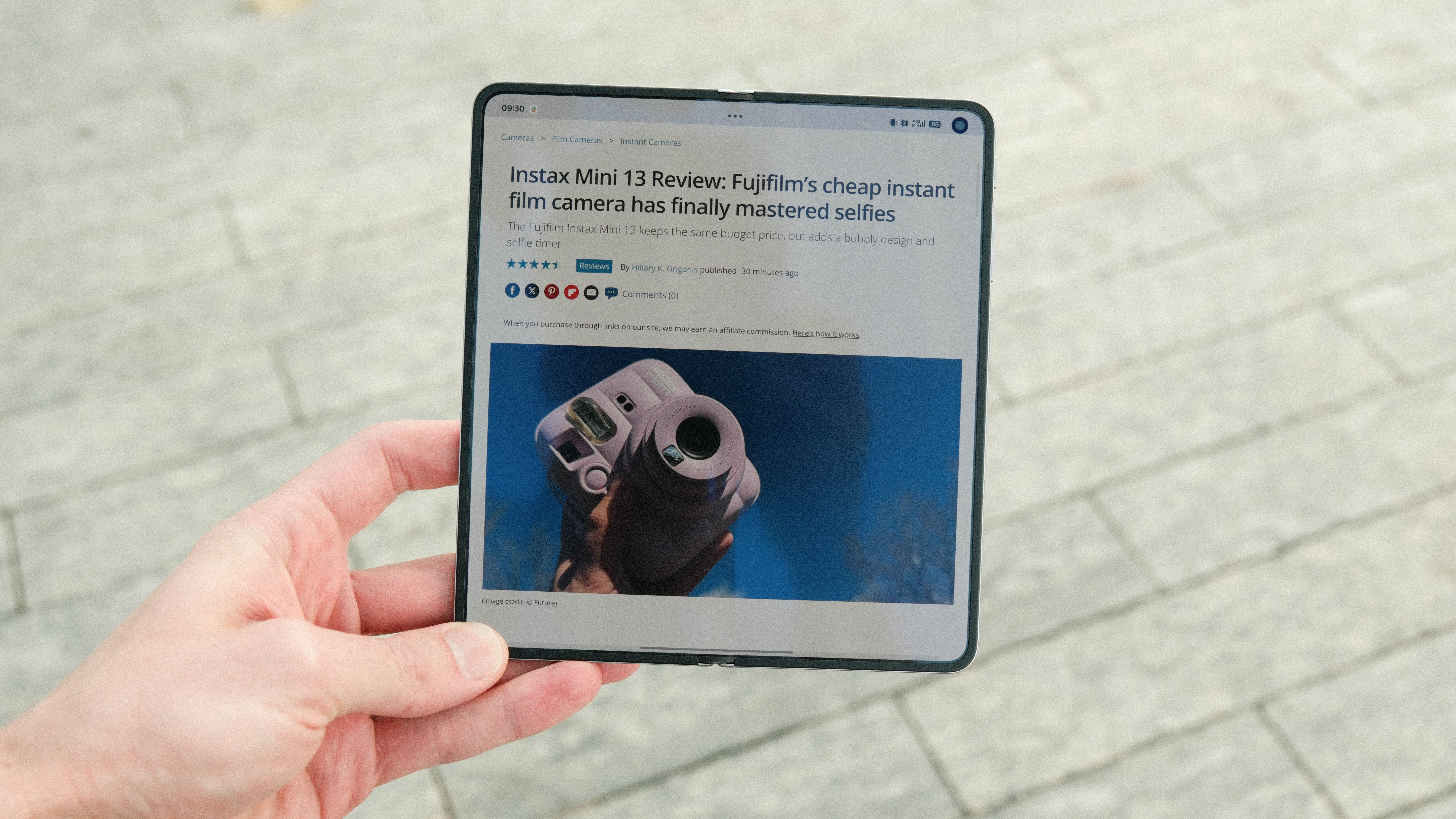 A person holding the device fully unfolded to reveal the large internal display. The screen shows a web browser featuring a review of a "Fujifilm Instax Mini 13" camera with a photo of the pink instant camera.