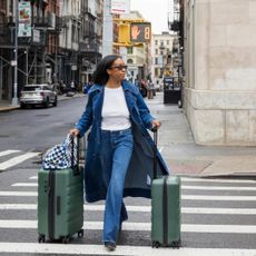 Woman in denim trench and jeans rolling green suitcases on a New York City street