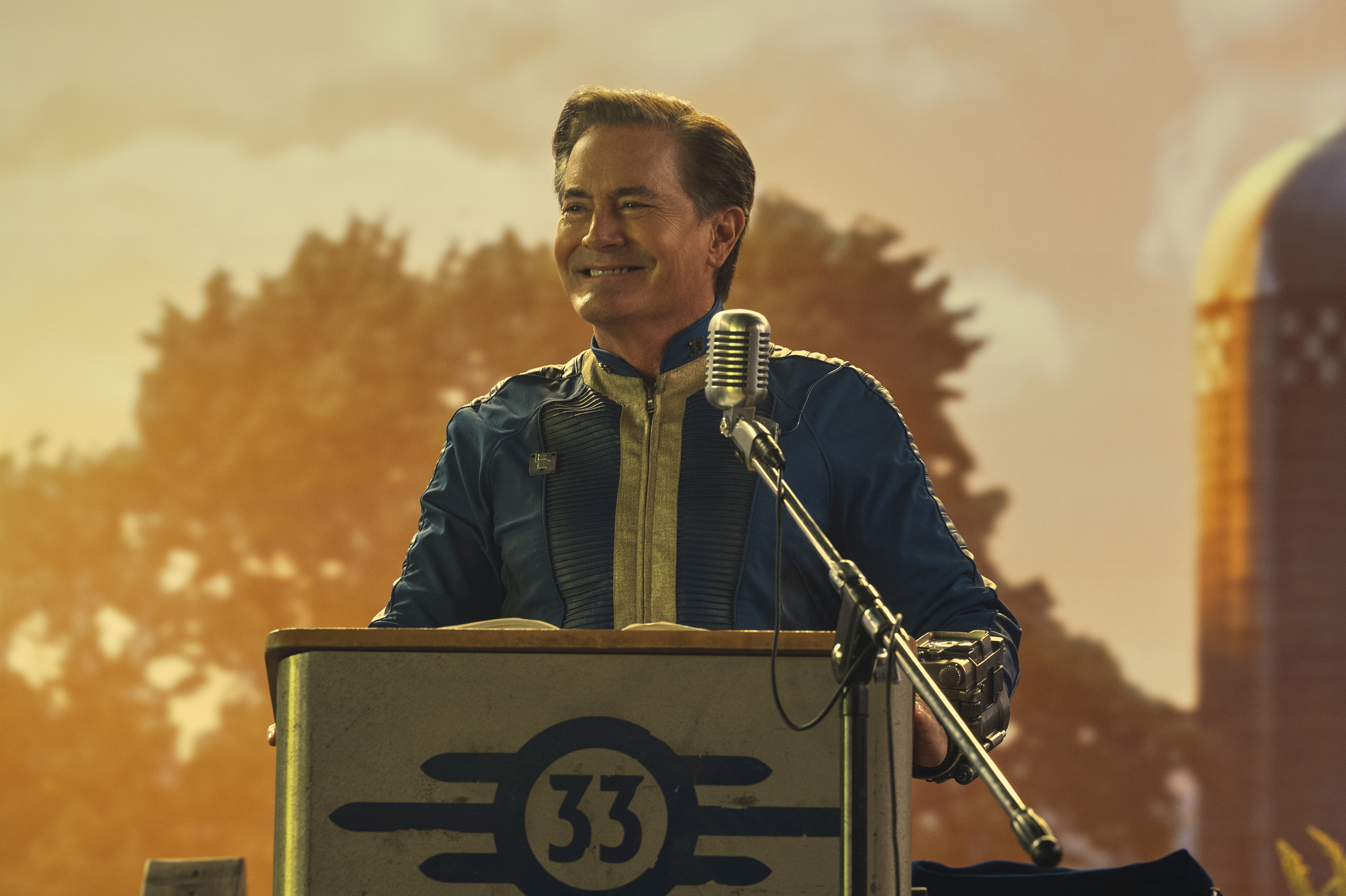 kyle maclachlan, standing at a podium with a microphone, in front of a projection of trees and a grain sill, in 'fallout'
