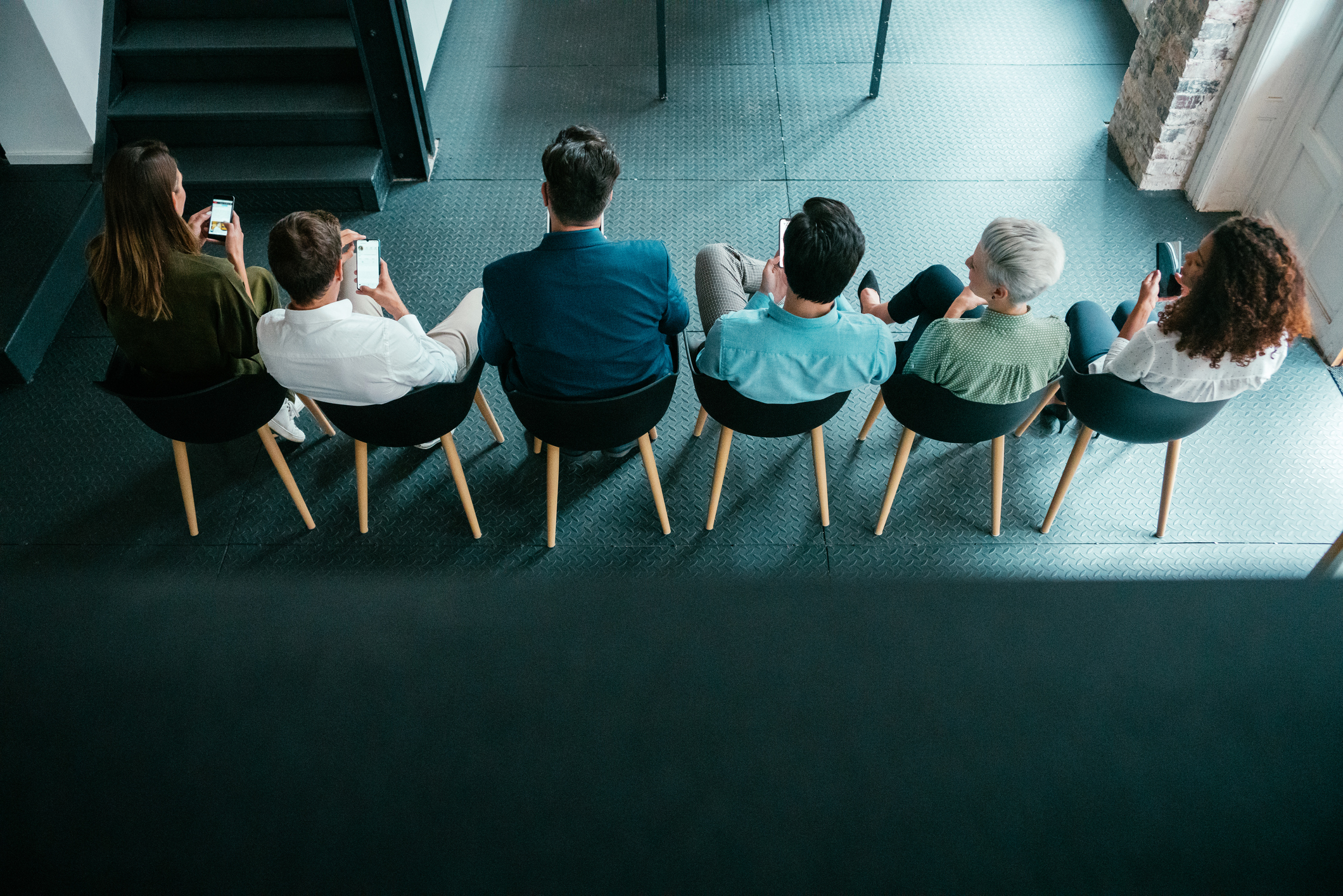 Rear view of a group of businesspeople sitting in chairs in the office and using their smartphones.