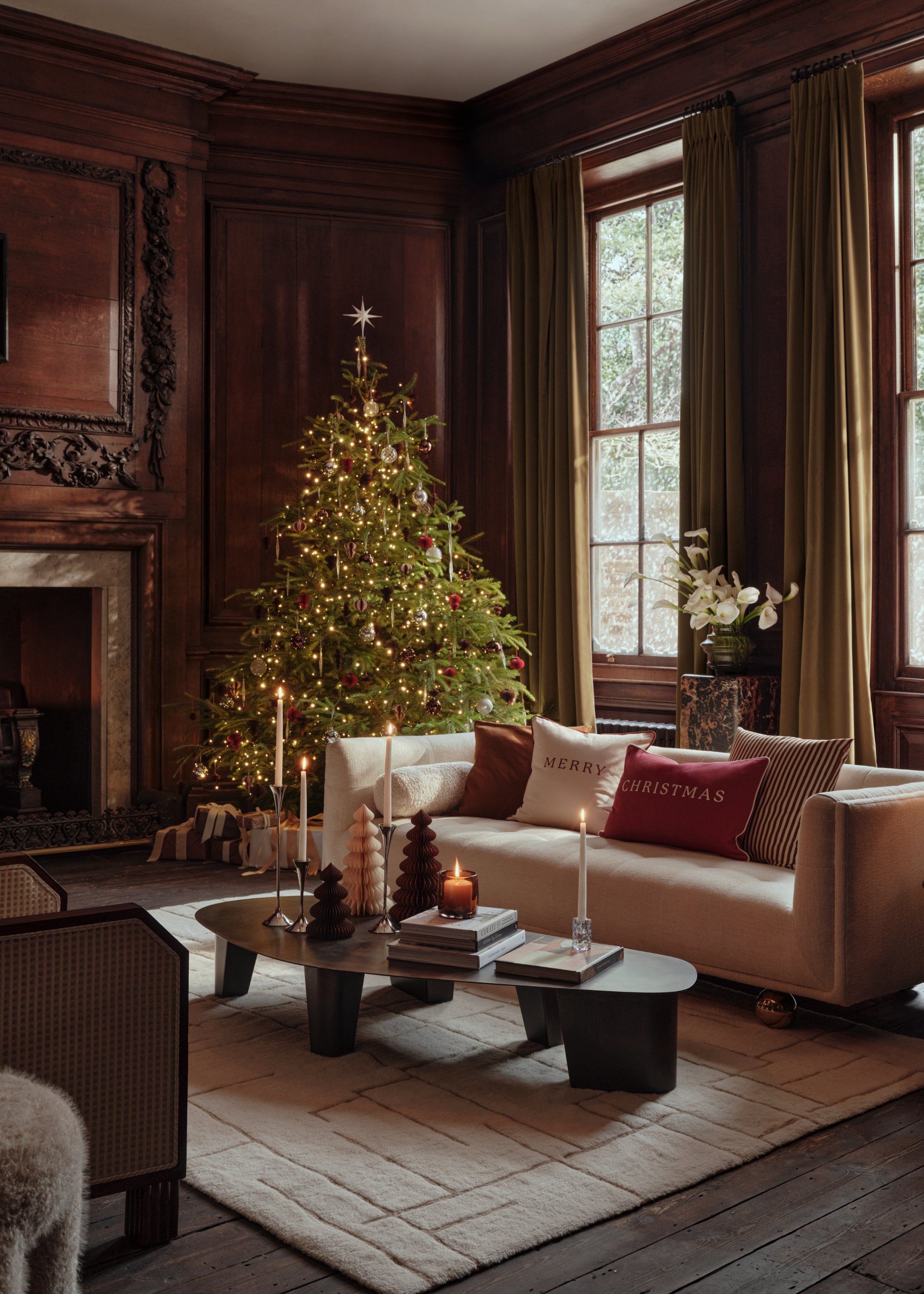 A Christmas living room with a large tree in the corner and lit taper candles at carying heights on the sculptural coffee table in the center with paper tree Christmas tree decorations