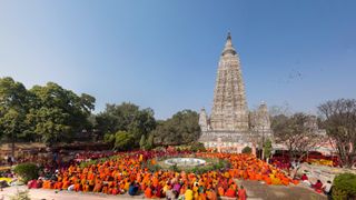 Hundreds of monks dressed in orange robes are praying in front of the Mahabodhi Temple in India. It is a very stall structure, with two smaller pillars either side.