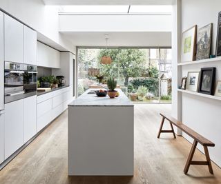 Airy and angular kitchen with kitchen island in white, wood flooring looking out on garden with French doors, and art work on shelves with bench below