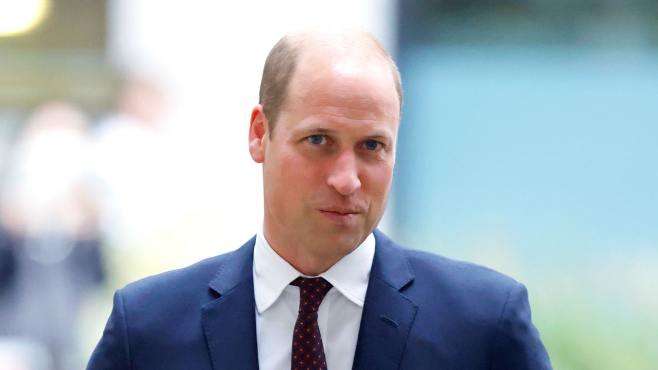 Prince William wears a blue suit with a white shirt and red tie as he smirks at the camera