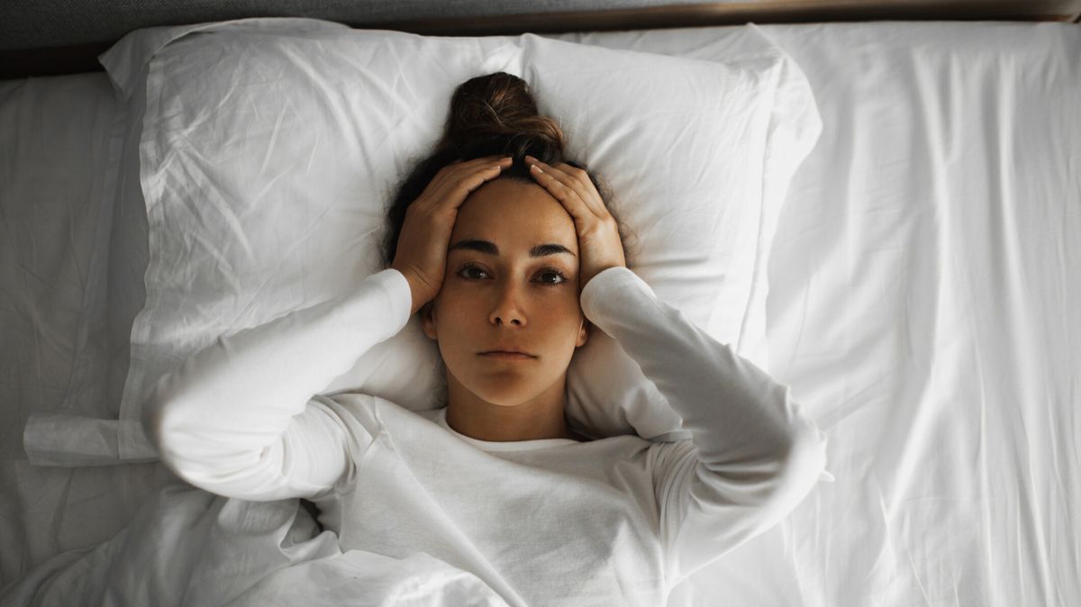 A woman with dark hair piled on top of her head lies on her back with her hands on either side of her face because she&#039;s wide awake at 3am and can&#039;t get back to sleep