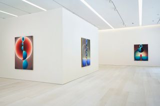 Three abstract pieces of art are hung in the all-white gallery room. The art is done in strong colors of blue and red, on a darker background.