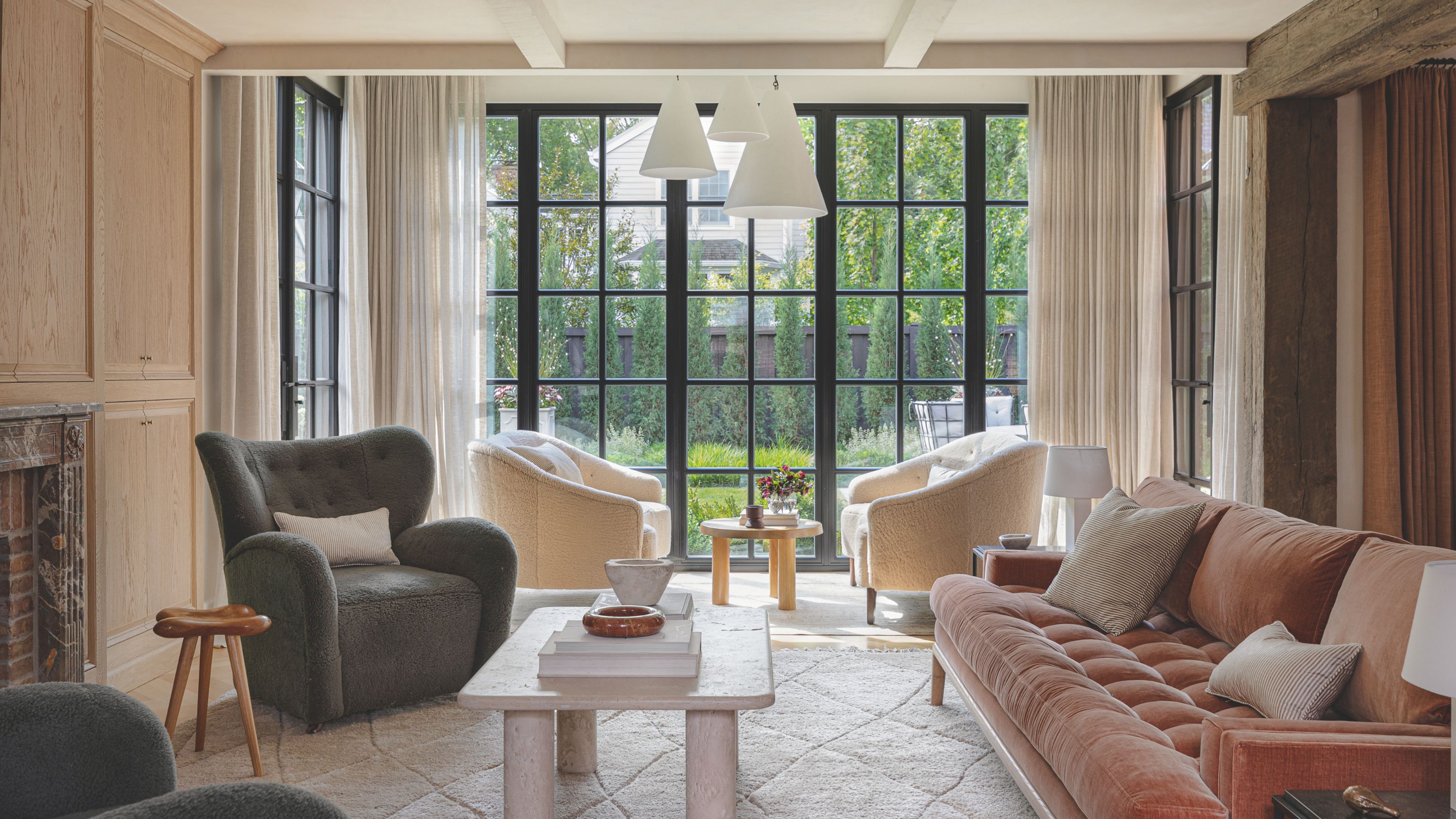 A bright, contemporary living room with floor-to-ceiling black-framed windows overlooking a lush garden. The space is furnished with a plush salmon-colored velvet sofa, textured armchairs, and a minimalist stone coffee table.