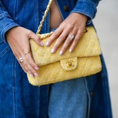 A tight crop of a woman wearing a double denim outfit, holding her hands (with pearlescent nails) over a yellow Chanel bag