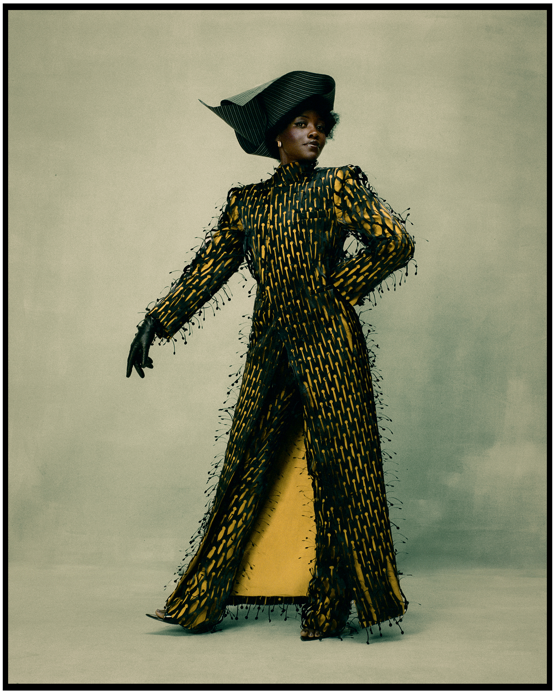 Lupita Nyong'o photographed by Kenny Germe&amp;#769; standing in front of a painted gray/blue backdrop wearing a black and yellow floorlength duster coat with matching pants and a black structured hat.