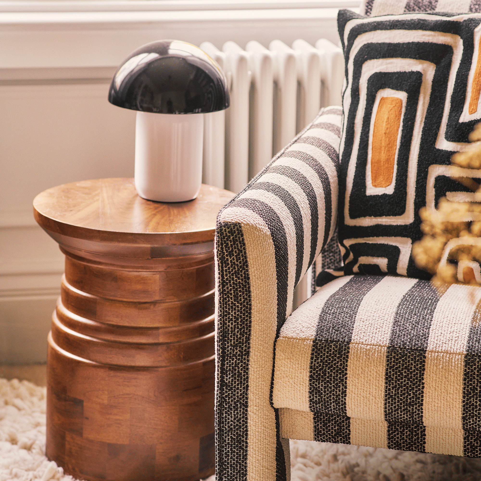 a living room with a close-up of a black and white striped armchair, a warm brown wood stool-style side table and a black and white mushroom-style table lamp
