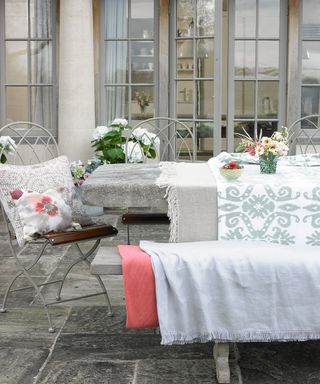 An example of outdoor dining ideas showing a dining table on a patio with grey chairs and blankets in shades of grey and pink