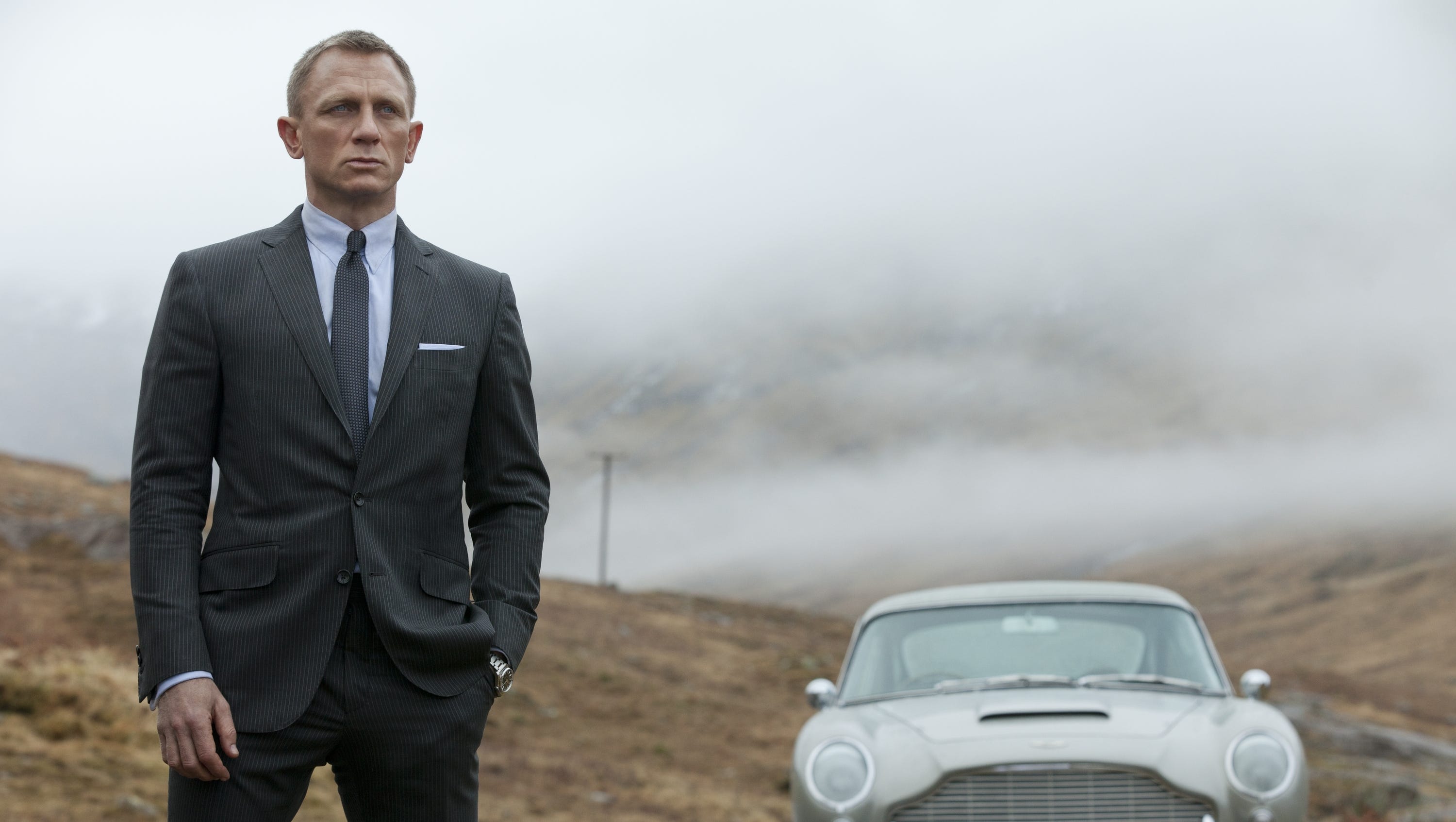 Daniel Craig as James Bond standing next to his Aston Martin in Skyfall