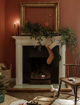 A living room with earthy terracotta wall decor, brass frame above Christmas-themed fireplace, wooden chair and logs stored in woven basket