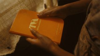 Image from the Marvel T.V. show Loki, season 2 episode 3. Image from the Marvel T.V. show Loki, season 2 episode 3. Close up of a man's hands holding a hardback orange notebook which has 'TVA Official Handbook' stamped in gold on the front cover.