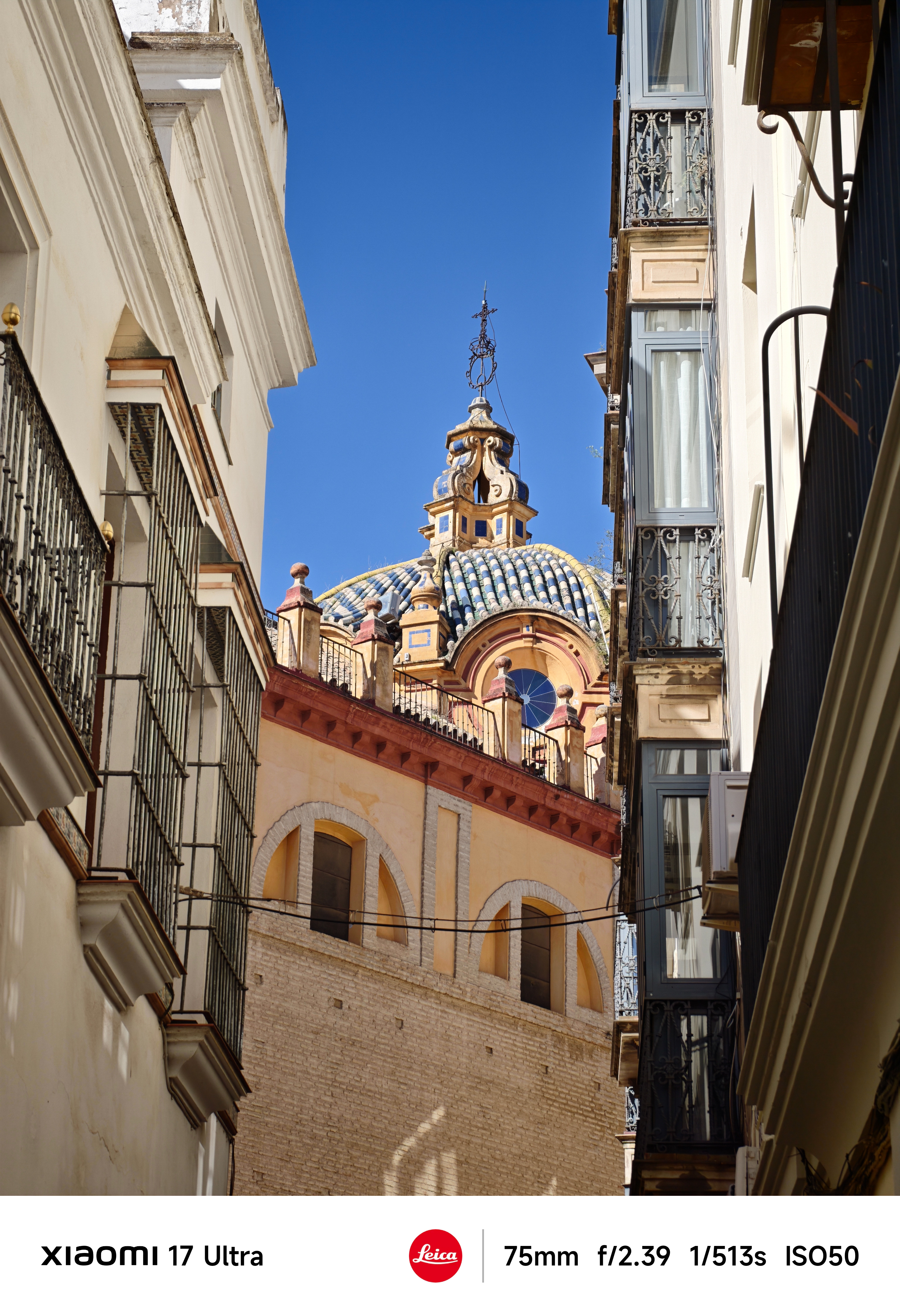 Decorative tiled dome framed between buildings, topped with a cross and ornate spire against a deep blue sky.