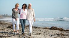 Three smiling older women walk arm in arm on the beach.