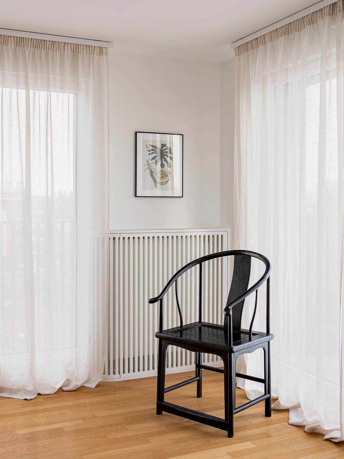 a white slatted radiator cover is used in the corner of a white room with wood flooring, white sheer curtains closed over the windows; there is a picture of a plant above the cover and a black armchair sits in front of it