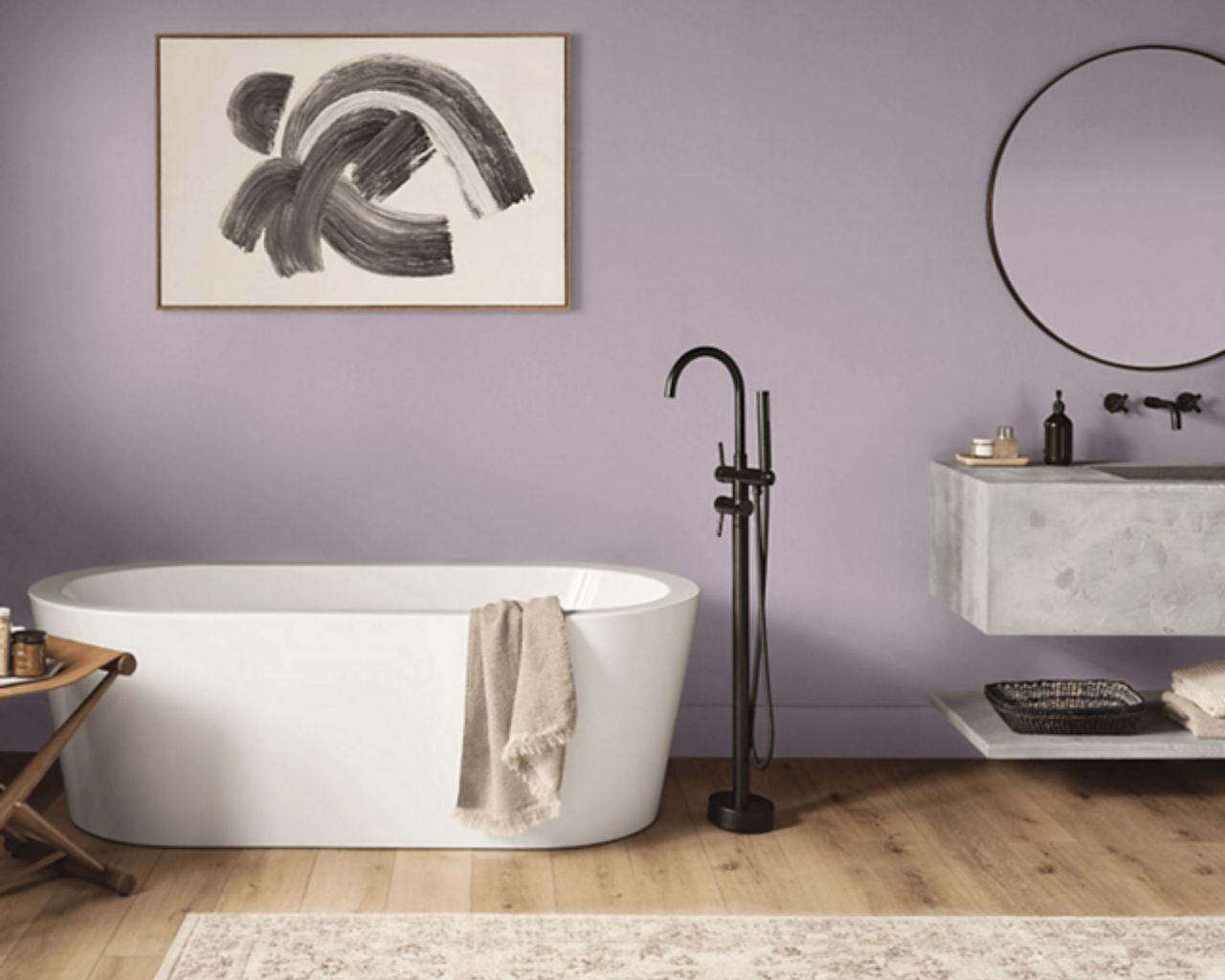 Mauve paint on a bathroom wall with art and a round metal mirror, white freestanding tub and marble wall vanity sink unit with floating shelf. Wooden floors and neutral rug for warmth