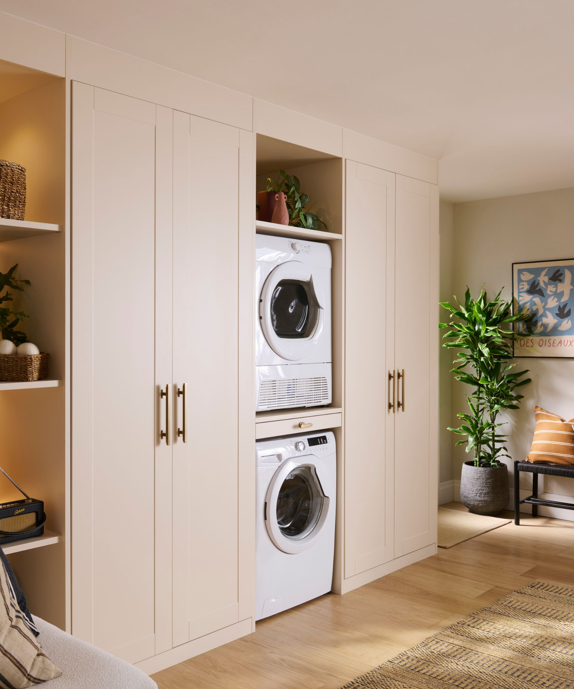 A wall of cream shaker cabinetry with brass handles conceals a stacked washing machine and dryer in a central open alcove, blending seamlessly into a living space with oak flooring.
