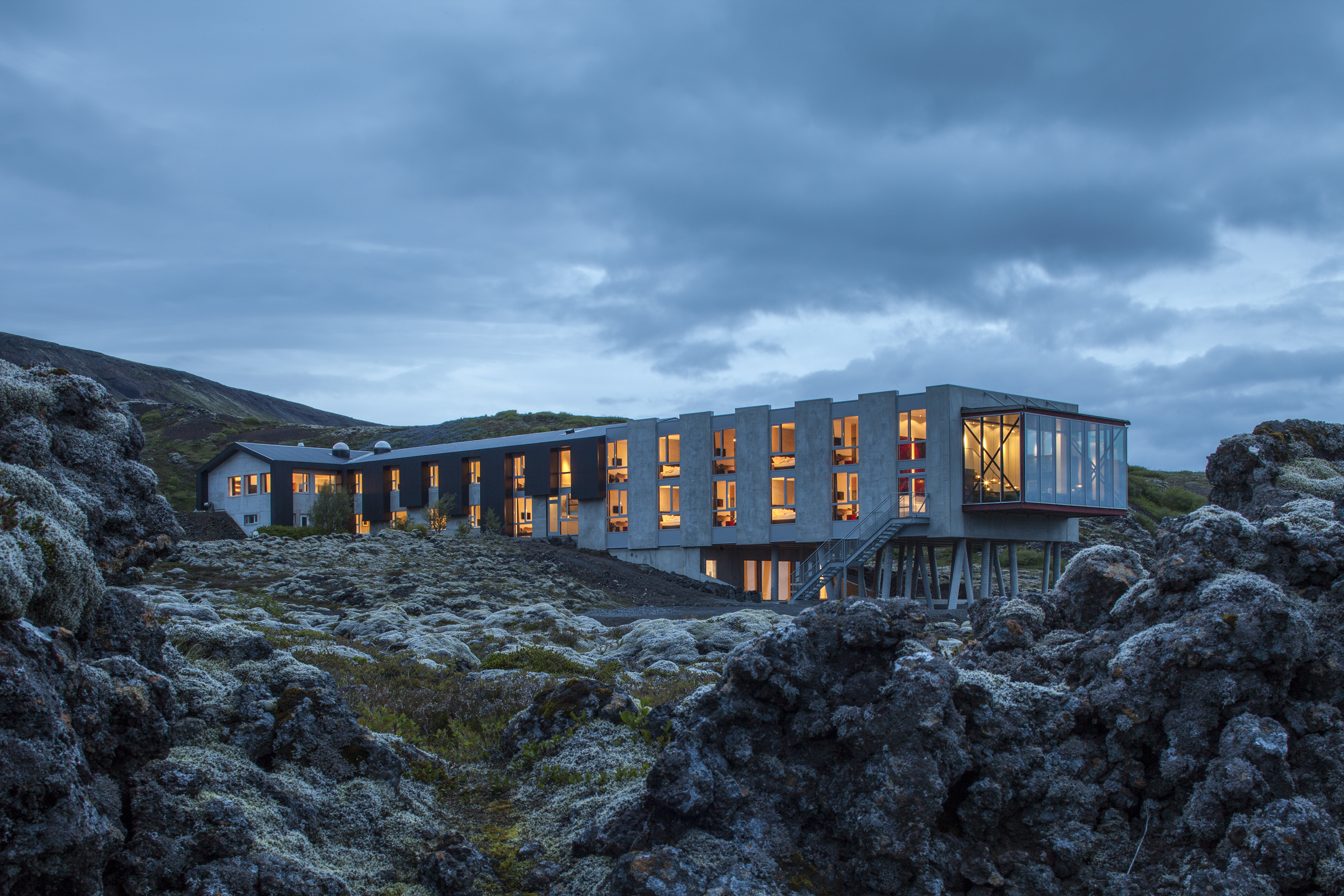 A hotel is nestled atop rocky formations in an Icelandic desert landscape, with floor-to-ceiling windows and minimal furnishings letting the outdoors in.