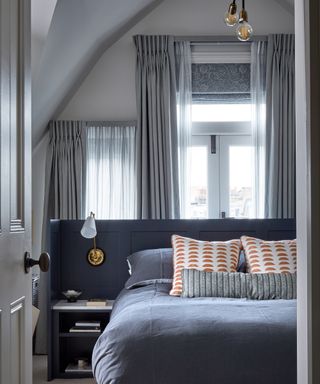 A bedroom with a blue bed and windows with added sheer curtains and a patterned blind