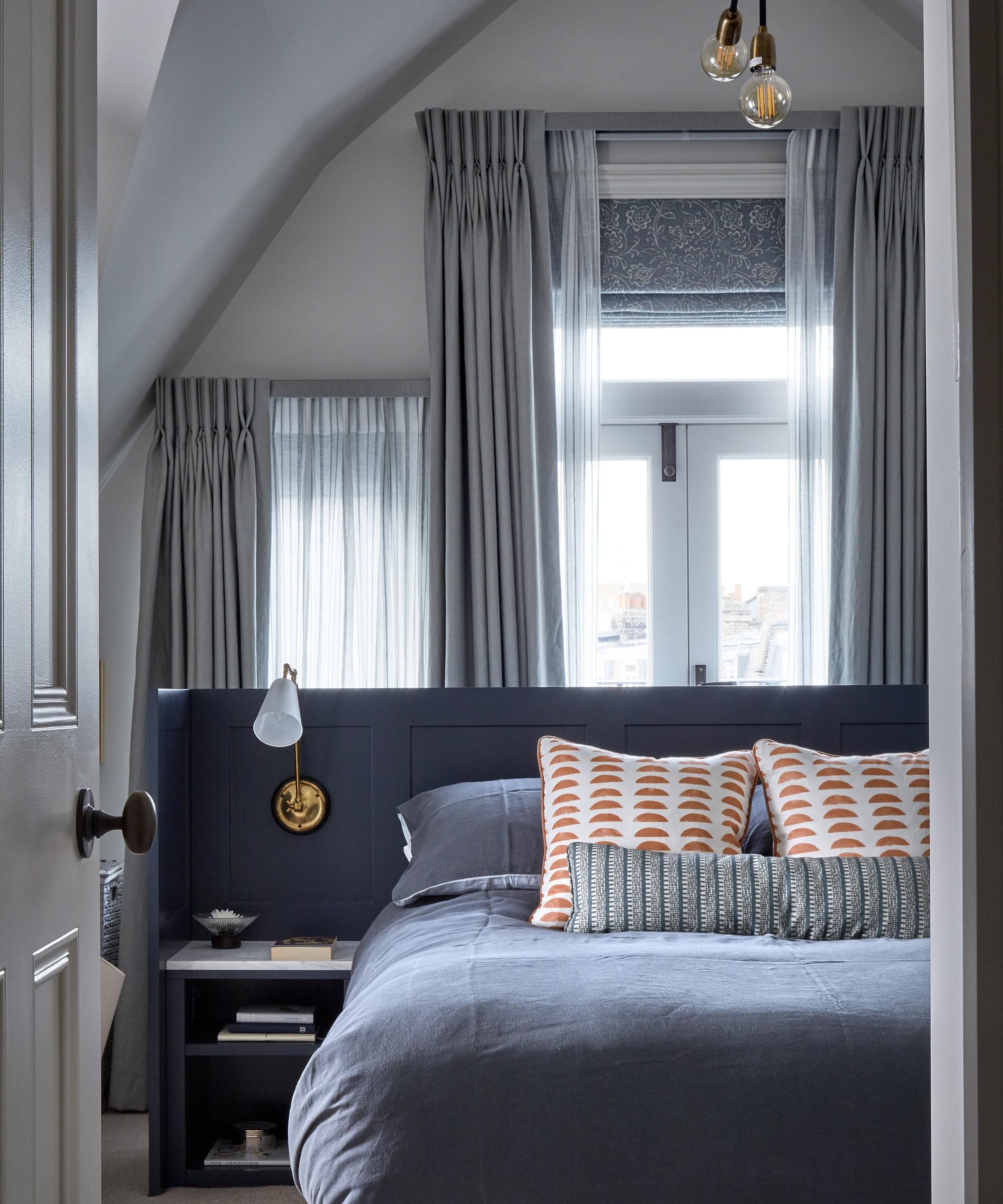 A bedroom with a blue bed and windows with added sheer curtains and a patterned blind