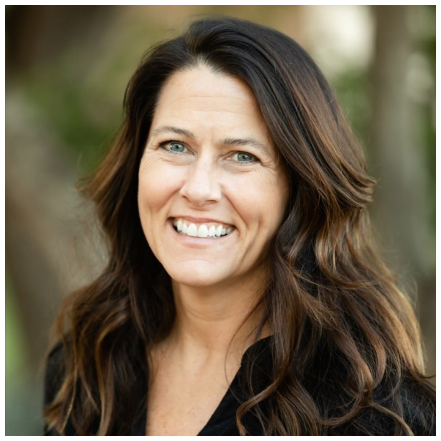A headshot of a woman with wavy brown hair loosely hanging over her shoulders, in a black v-neck top, smiling
