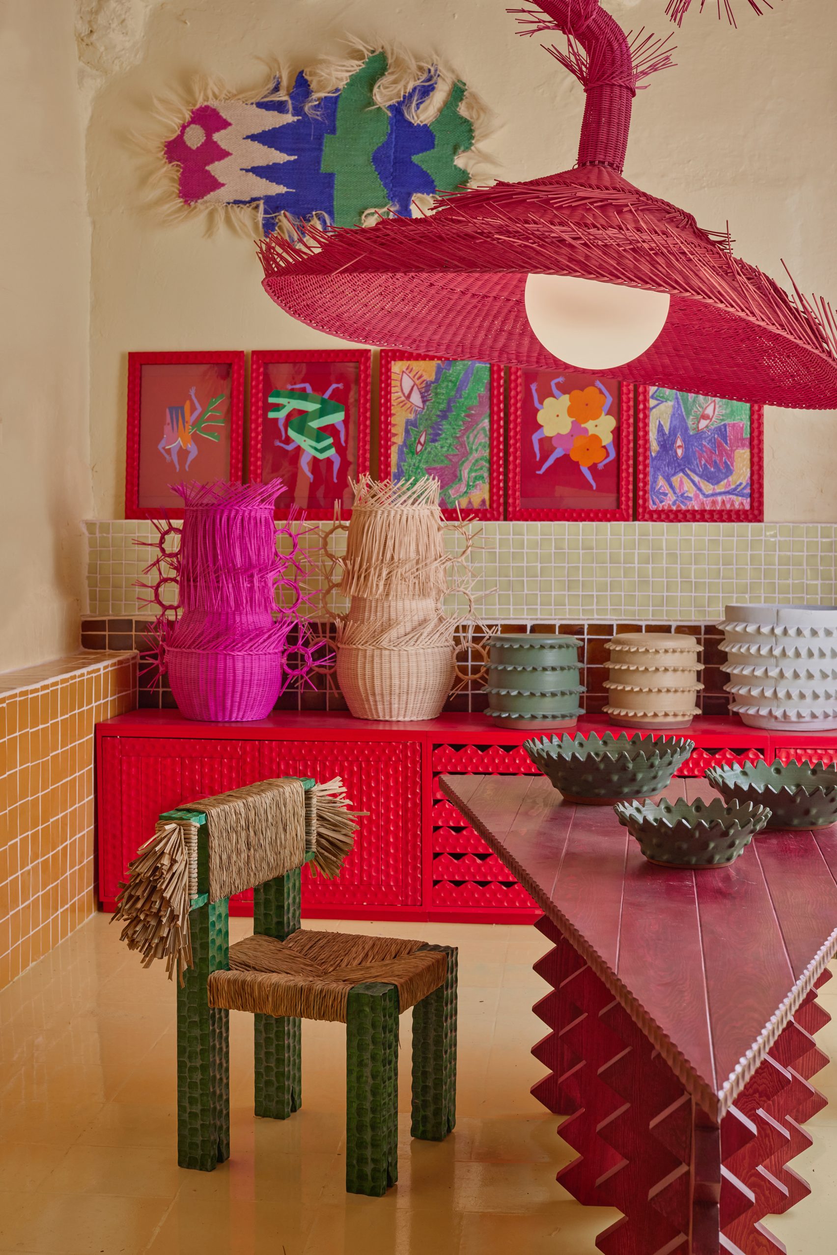 An irreverent design studio with geometrically cut rattan creations in bright pink, natural, and bordeaux, a triangular table with zig-zag legs in red, ochre tiles, and colorful prints.