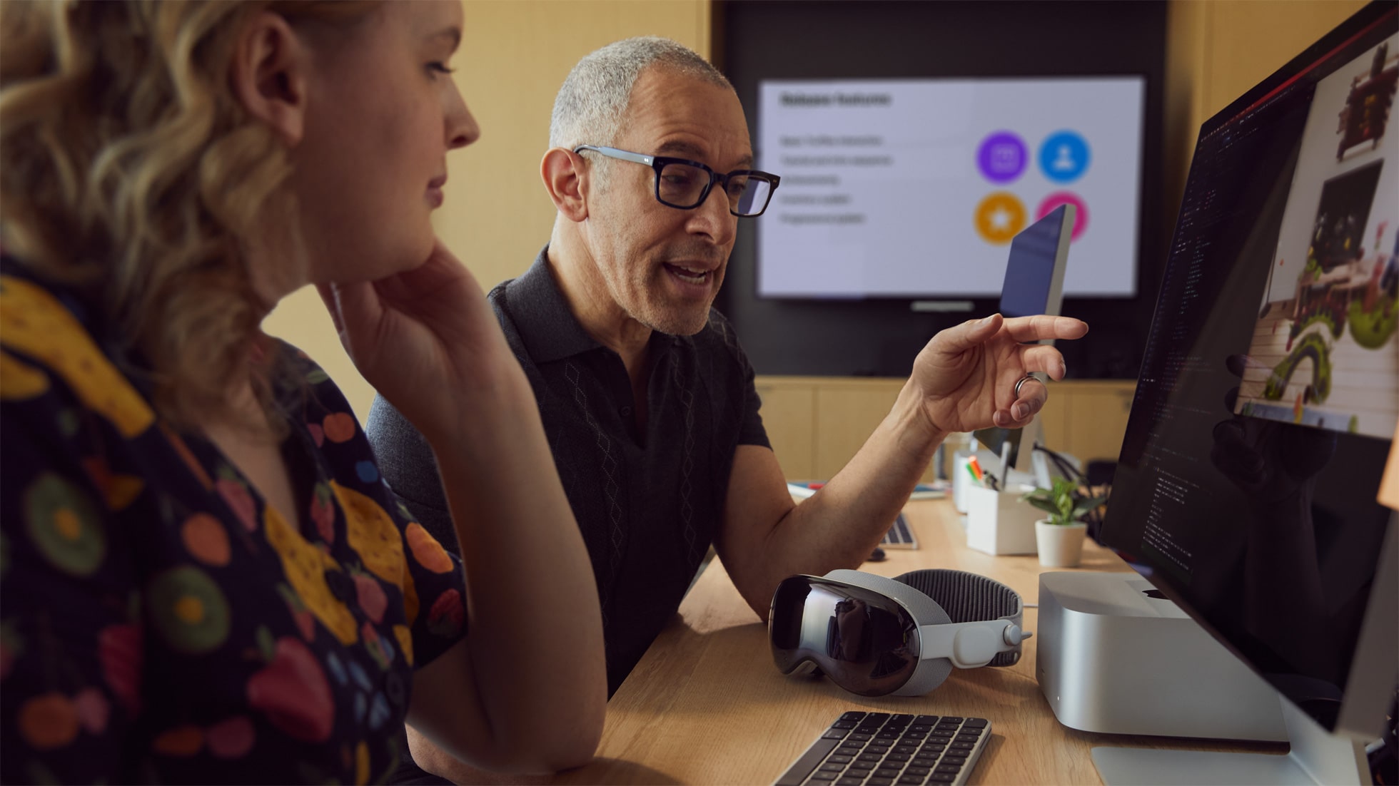 Dos personas se sientan en un escritorio con un Mac Studio, un Studio Display y unos auriculares Vision Pro frente a ellos.