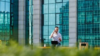 A man sat on a bench on the phone, with a laptop on his lap and a coffee sat next to him. It is a sunny day and he&#039;s sat in what looks like a business park, partly obscured by shrubs in the foreground, which are out of focus. It&#039;s ambiguous whether or not the man is working remotely.