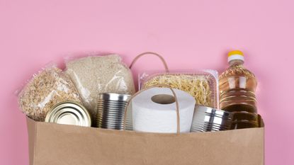 Paper bag with crisis food supply for the period of quarantine isolation on pink background with copy space, rice, pasta, oatmeal, canned food, toilet paper. Food delivery, donation during coronavirus.