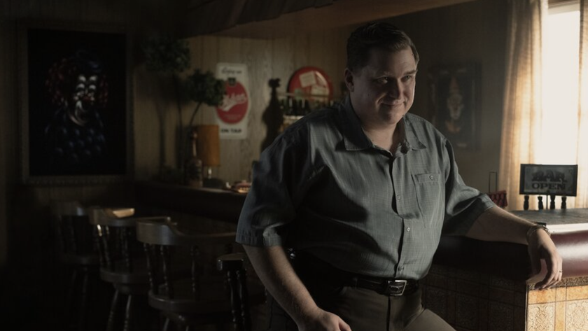 John Wayne Gacy (Michael Chernus) perched at a bar, with pictures of clowns visible on the walls, in Devil in Disguise: John Wayne Gacy