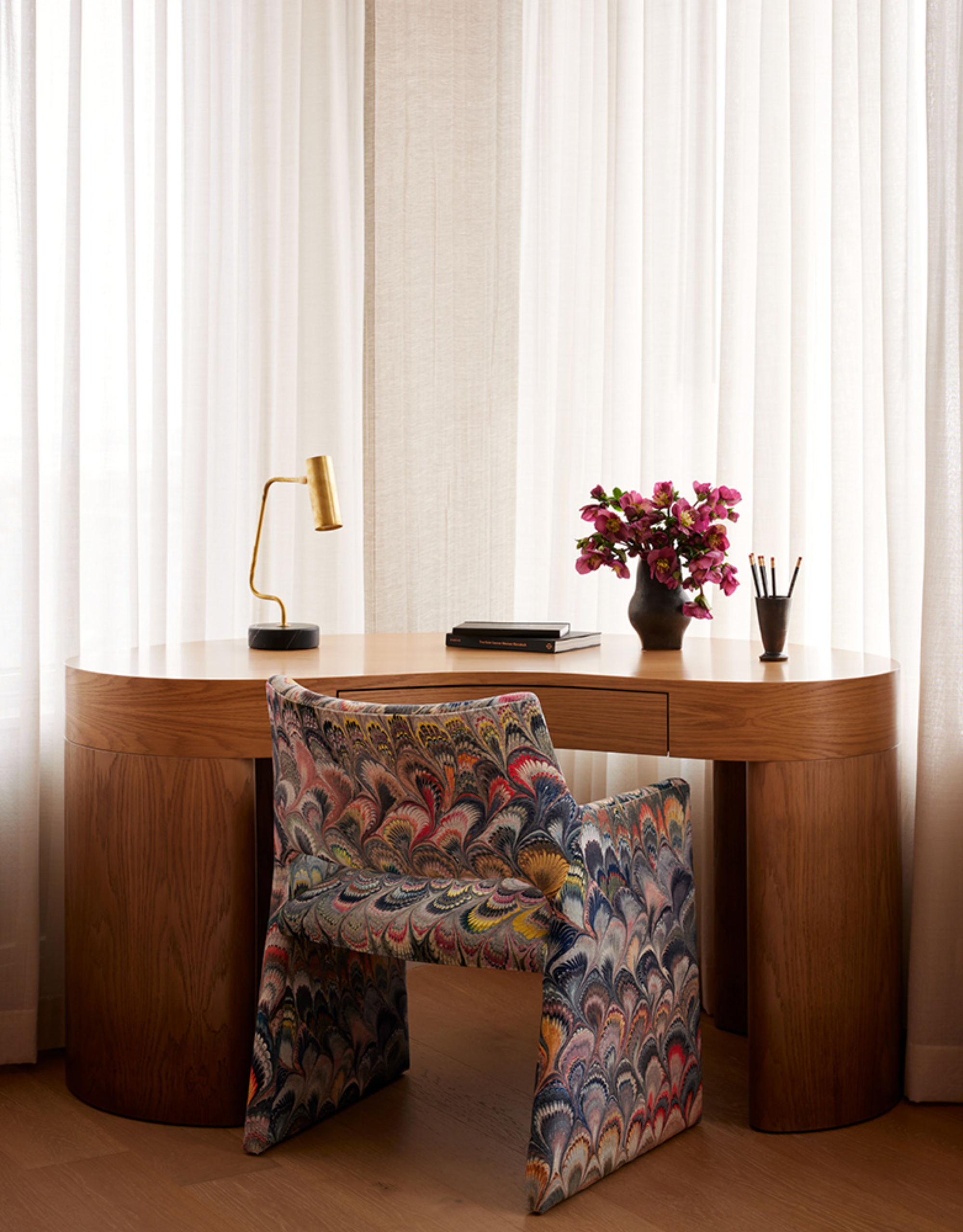 A wooden bean-shaped desk is surrounded by sheer curtains and a marbled armchair. On the desk is a gold table lamp, a vase of purple flowers, a pen holder, and two stacked books