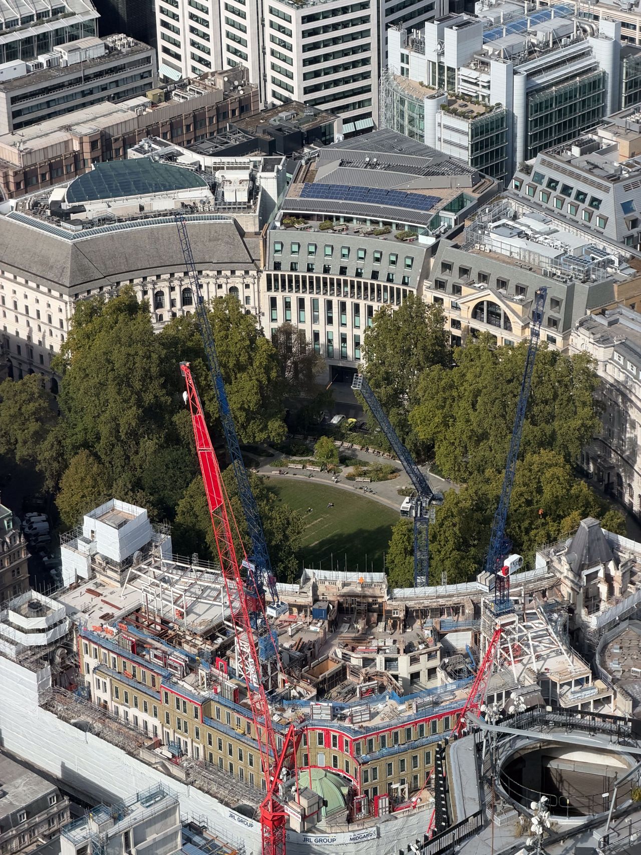 Aerial view of a large central London construction site with multiple cranes beside a tree-filled garden and office buildings.