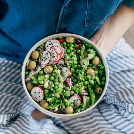 Fibremaxxing: A woman sat cross legged eating a bowl of vegetables, including peas, radish, olives, and salad leaves