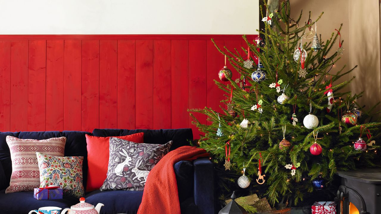 Decorating ideas for Christmas, foliage garlands on the ceiling, blue sofa in front of red painted panelling, cushions with floral patterns, stack of presents underneath a decorated Christmas tree