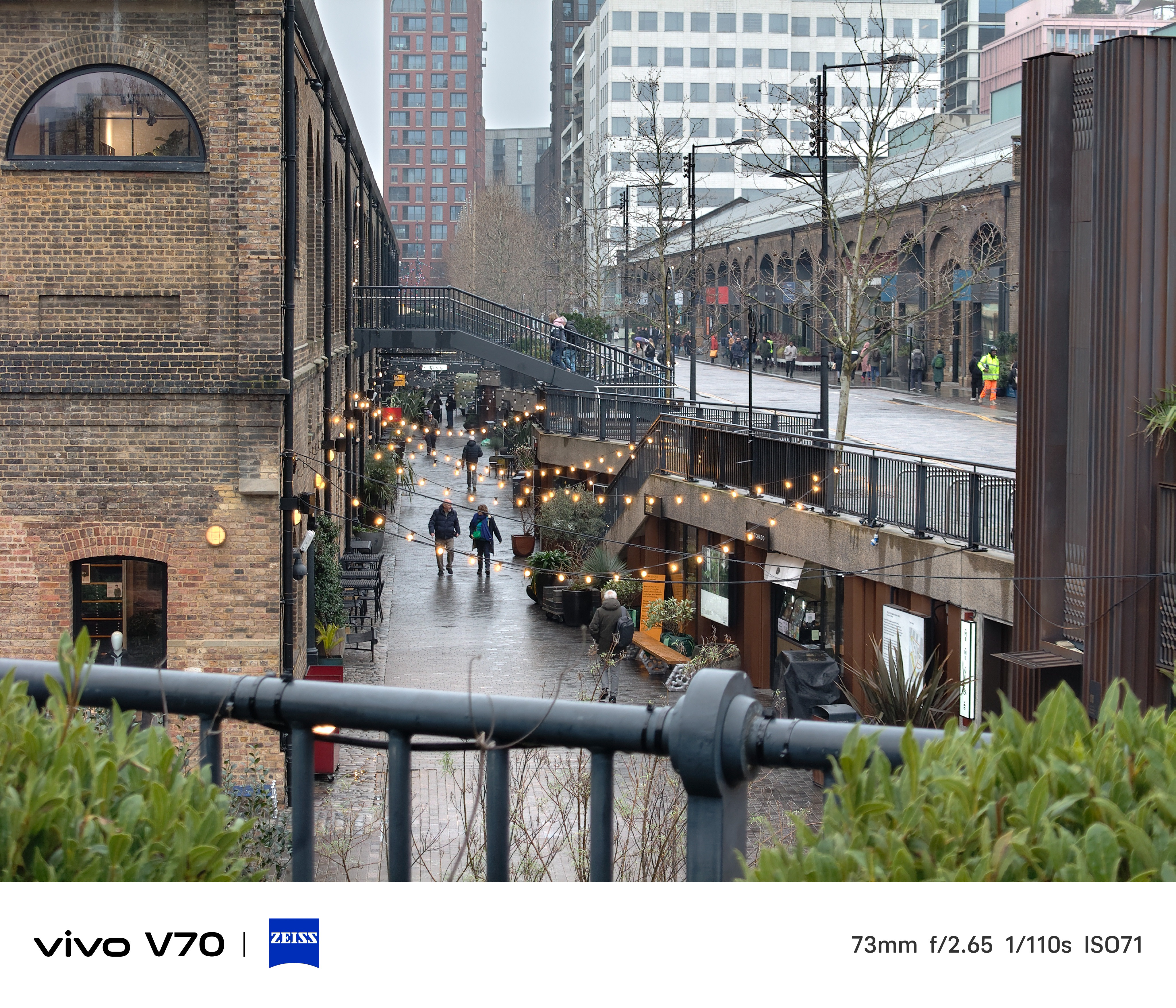 Rainy Coal Drops Yard walkway in London with string lights glowing between brick buildings and pedestrians below.