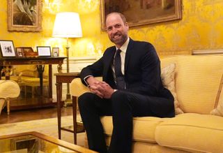 Prince William wears a suit while sitting on a yellow coach and he has a stubbly beard